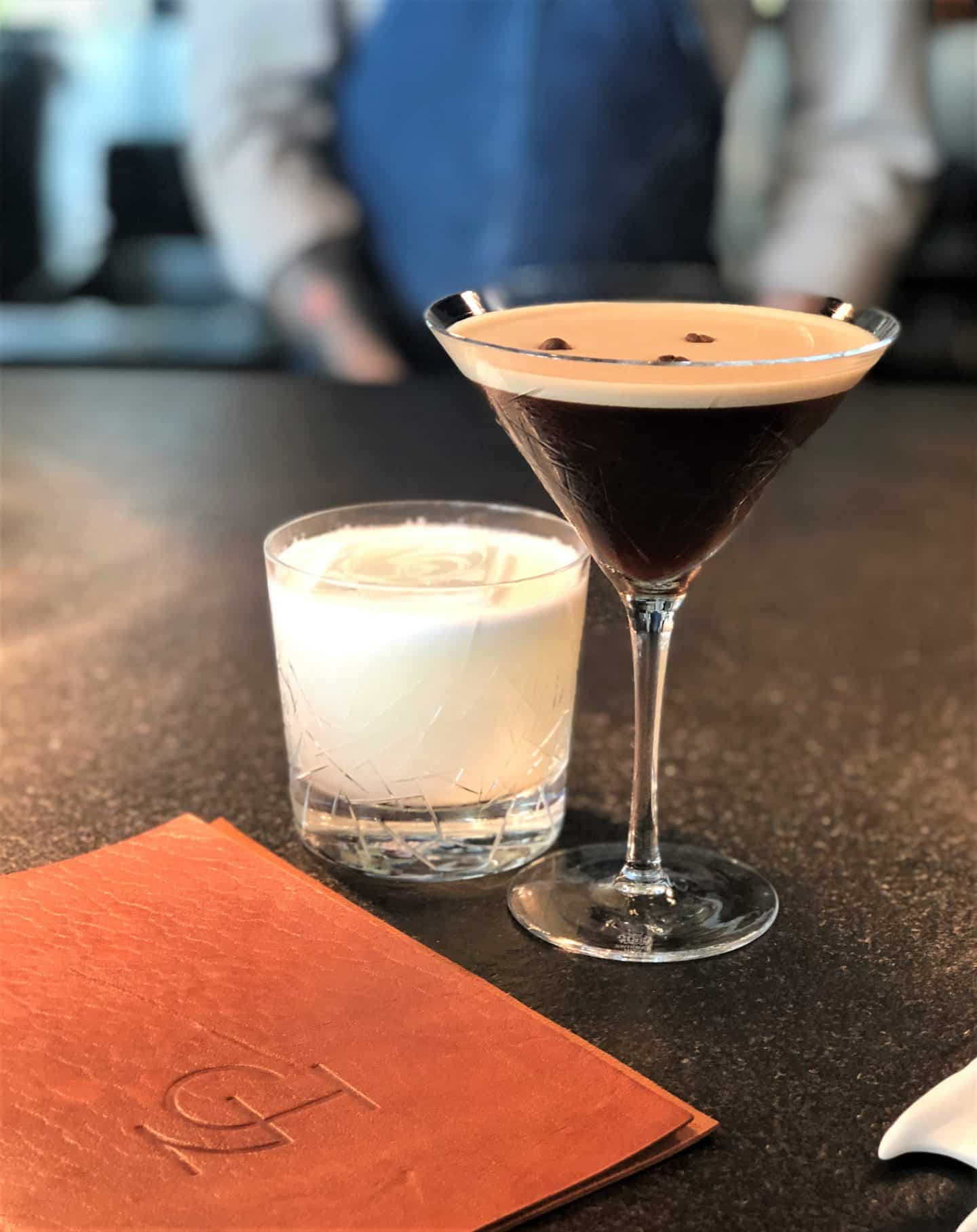 An Espresso Martini and Pisco Sour with the tan leather NOA mena on the black counter