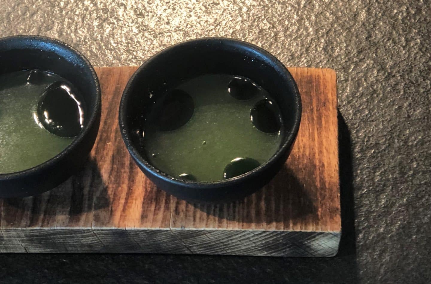 A black small on a wooden board. The bowl contains the fermented celeriac with apple cider balsamic vinegar and is green in colour Noa Chef's Hall, Tallinn