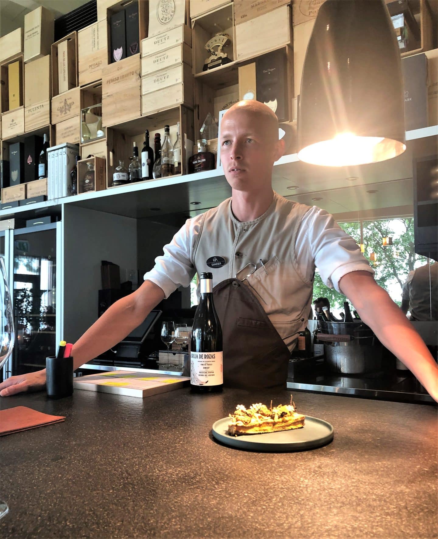 Sander the sommelier standing in front of one of the dining courses at the Chef's Hall in Tallinn with wines and spirits behind him.