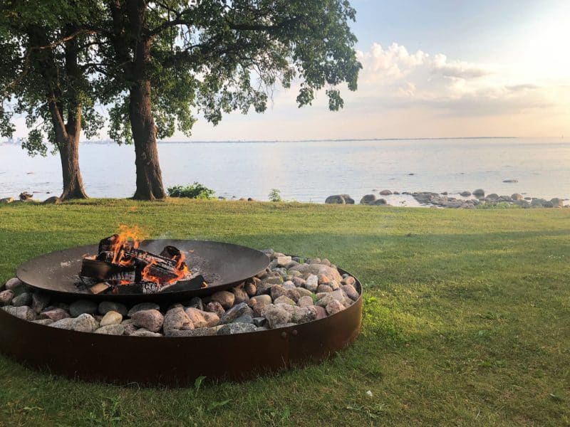 The fire pit at Noa Chef's Hall in Tallinn with Tallinn Bay in the background close to sunset time. Theer are trees to the side and grass surrounding the raised fire pit on large pebbles. 