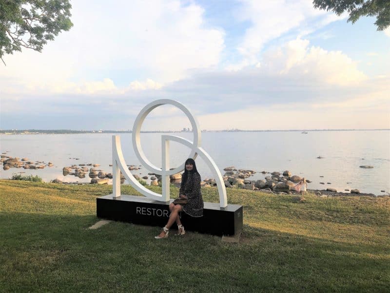 Bejal wearing a black knee length dress with pink flowers with Tallinn Bay in the background. She is sitting on the platform of the Noa Restorans stone logo, Tallinn before diningining at Noa Chefs Hall in Tallinn