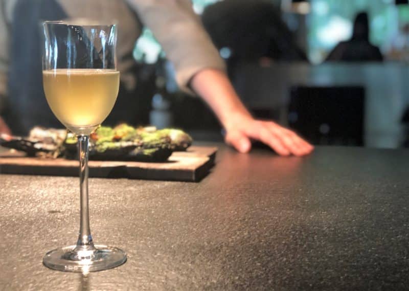 A glass of wine with Chef Roman in the background standing over a board of charred leeks and truffle at Noa Chef's Hall, Tallinn