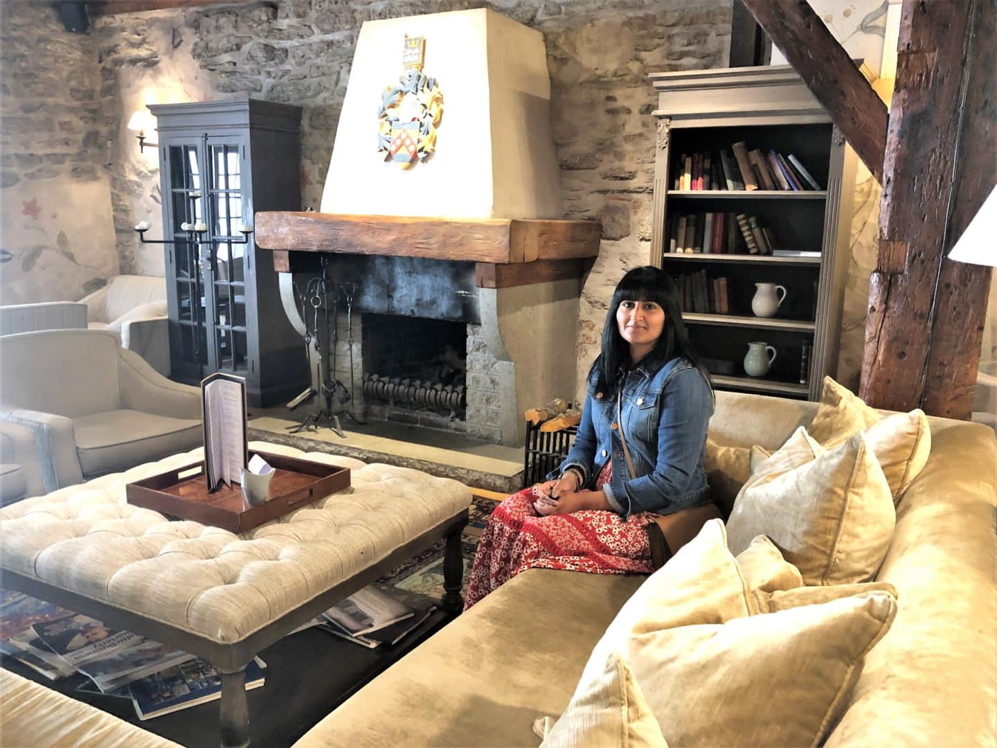 The Schlossle Hotel Tallinn: The lounge and lobby with beamed brown ceilings, a huge comfy corner sofa and stoned walls. bejal is sitting on the sofa wearing a red dress with denim jacket and there is an open fireplace behind here with a padded coffee table right infront of her.