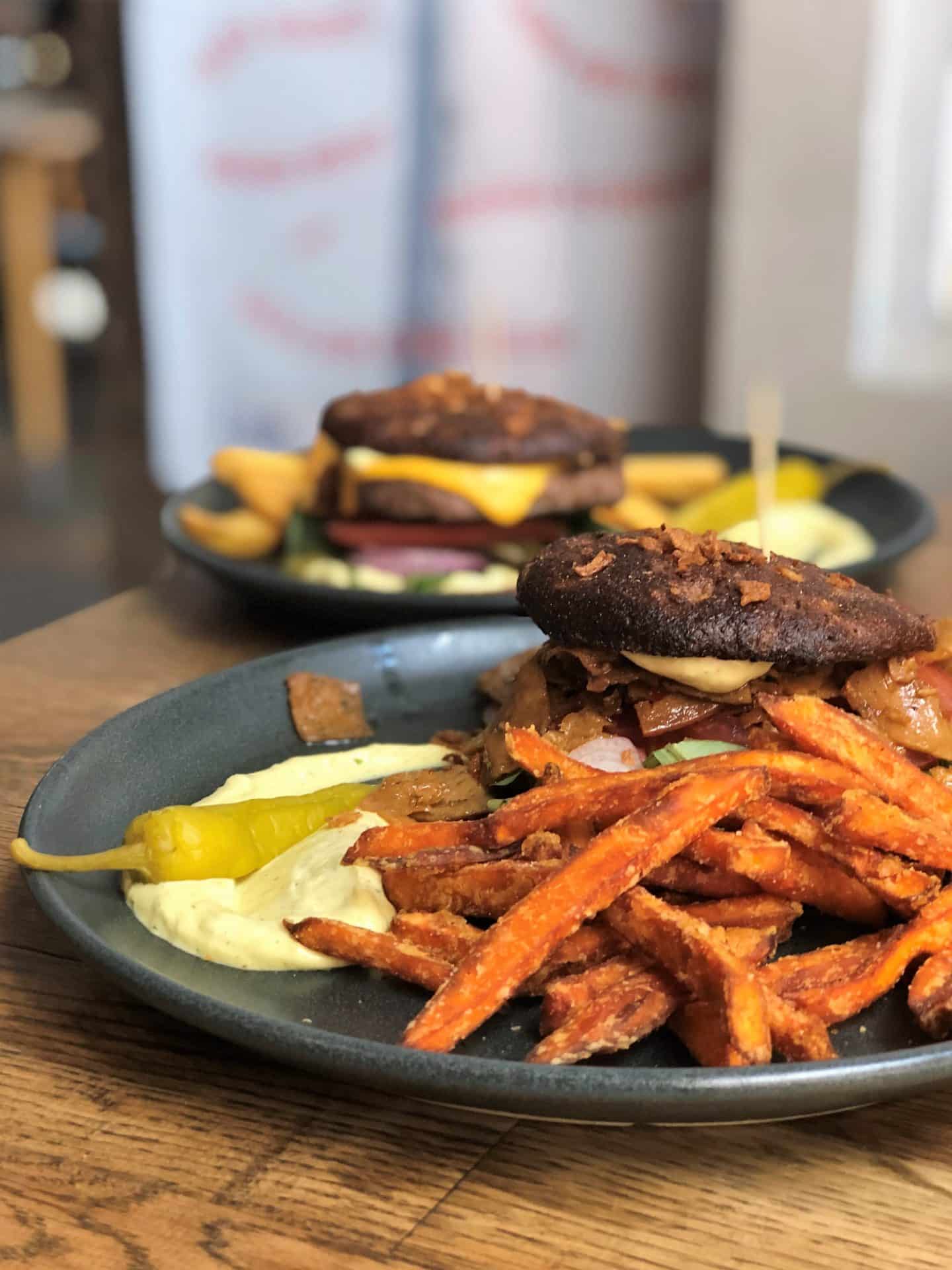 Side veiw of vegan Furberger which is a dark bread burger with beetroot hummus, parsley and chilli pepper aioli tomato and red onions and a side of fries on a dark wood table at F-Hoone, Tallinn