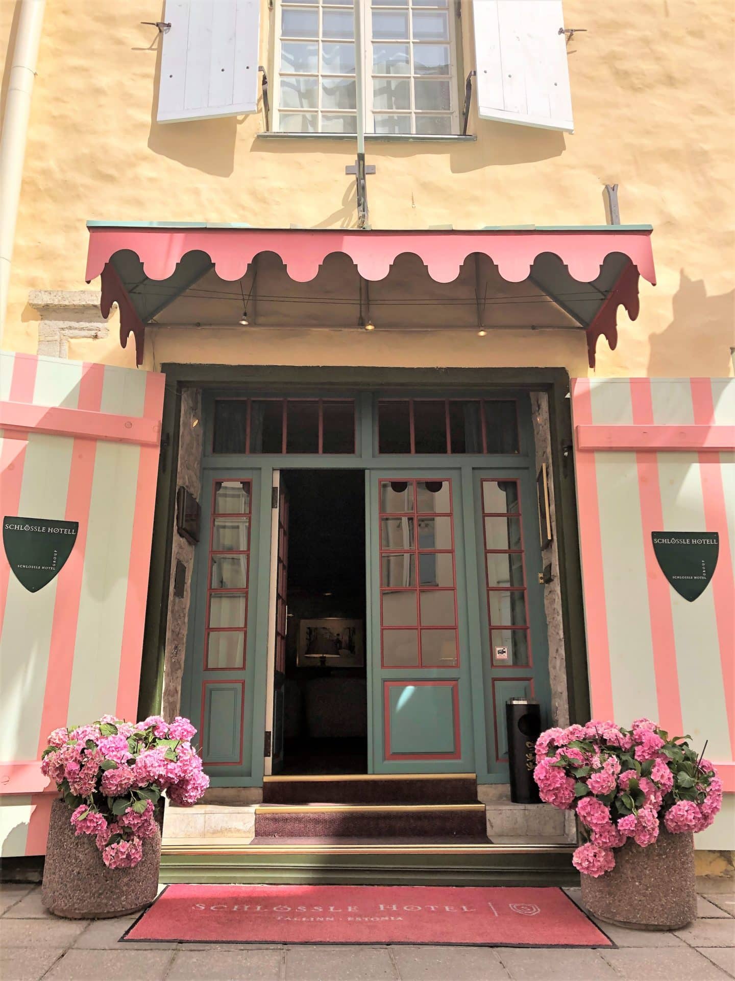 The Schlossle Hotel Tallinn pink and duck egg blue door with pink and pale green strip shutters. There are 2 pots of pink Hydrangea bunches at either end of the entrance