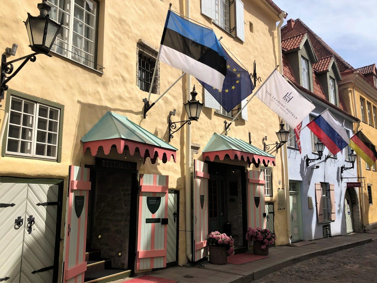 Schlossle Hotel Tallinn: A luxury Hotel Review. The exterior pf teh hotel with yellow walls, Estonian flags and pink adn pale green awning, on a cobbled stoned street.