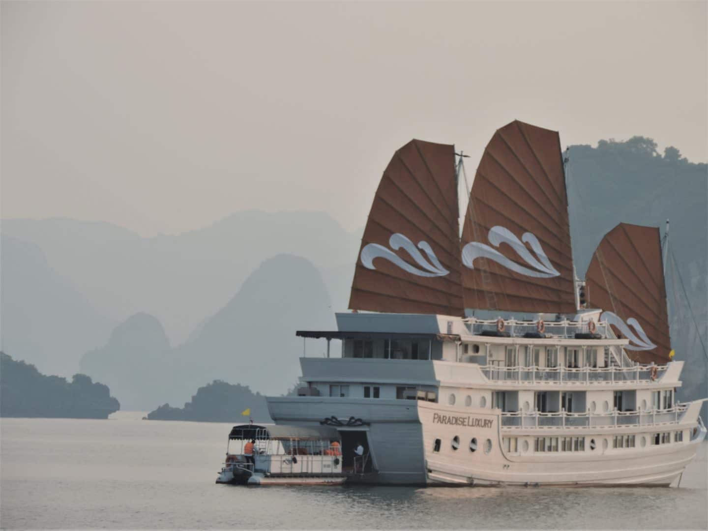 The Paradise Luxury cruise ship on Halong Bay with it's red-orange 3 sails, white exterior and the limestone formation of cliffs in teh background.
