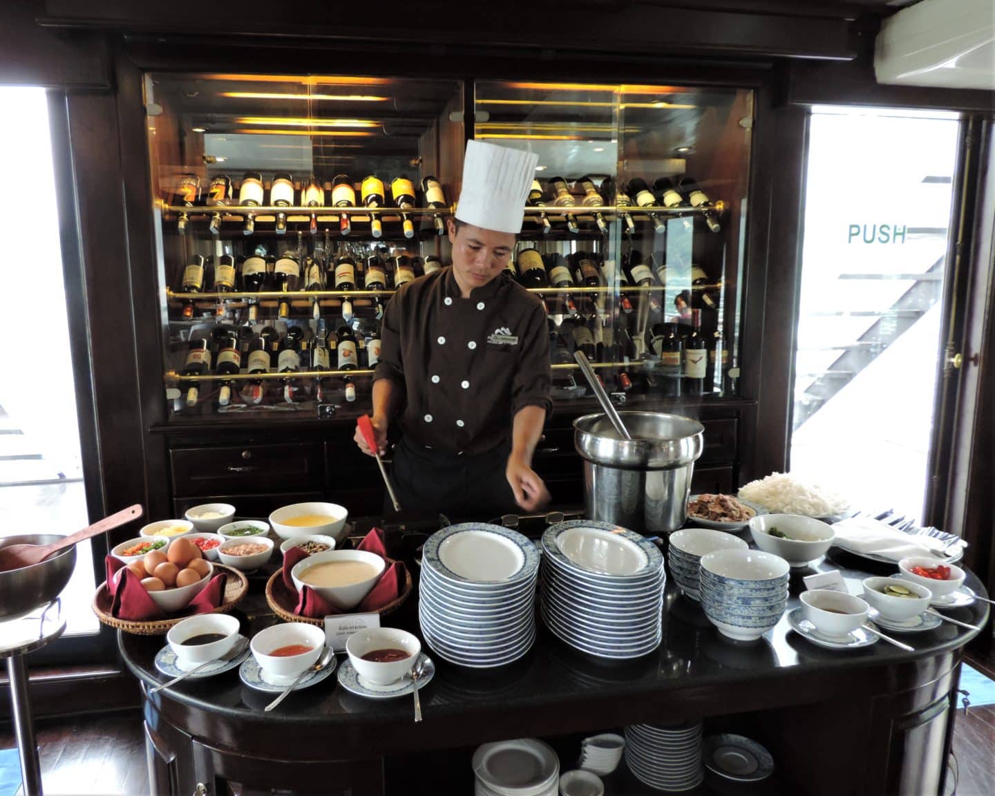 Halong Bay chef serving food aboard the Paradise Luxury. there are wine bottles in the background and a stack on white plates with bowls of sauces on the counter, Halong Bay.