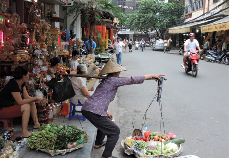 Vietnam in 14 Days: Lady carrying fruit and veg in Hanoi wearing a purple to and black trousers, with market vendours behind her and a man on a motorbike going past.