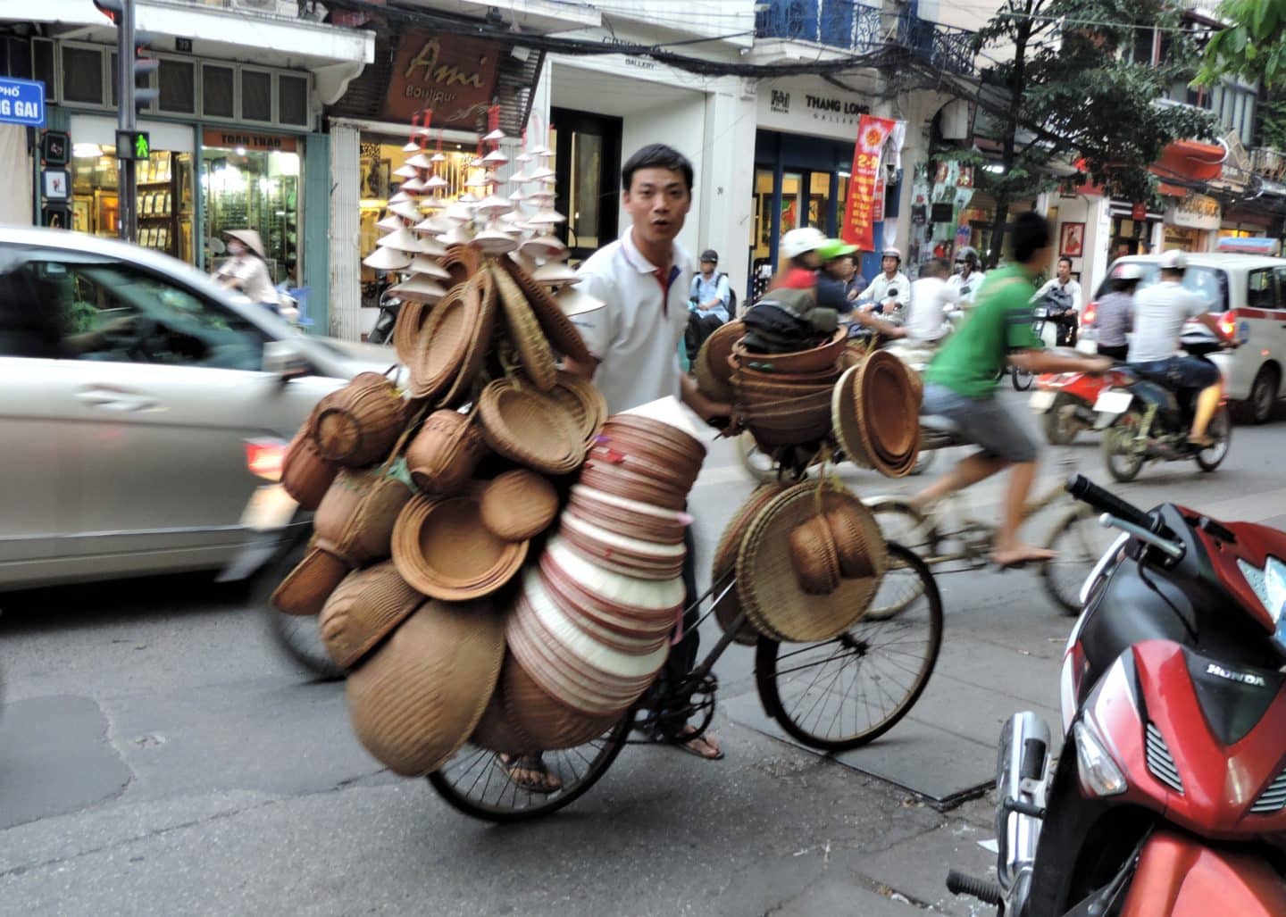 Man carrying hats on bike in Hanoi with cars and people walking by in the background.