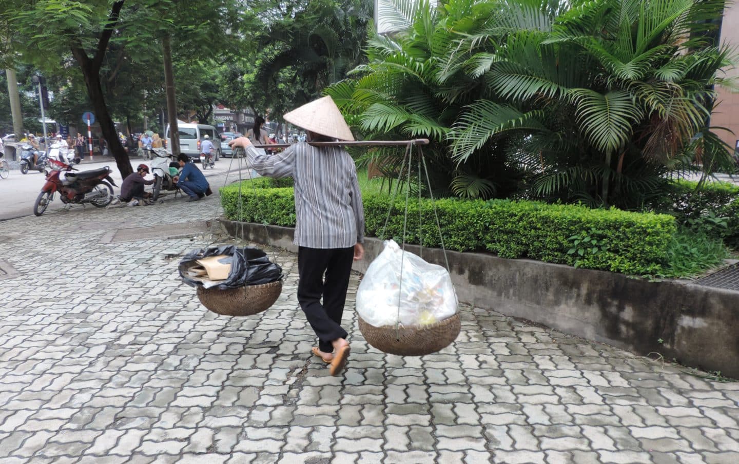 Lady carrying plastic in Hanoi, Vietnam on a bamboo pole. She is wearing the traditional Nón Lá hat and is walking passed a hedge.