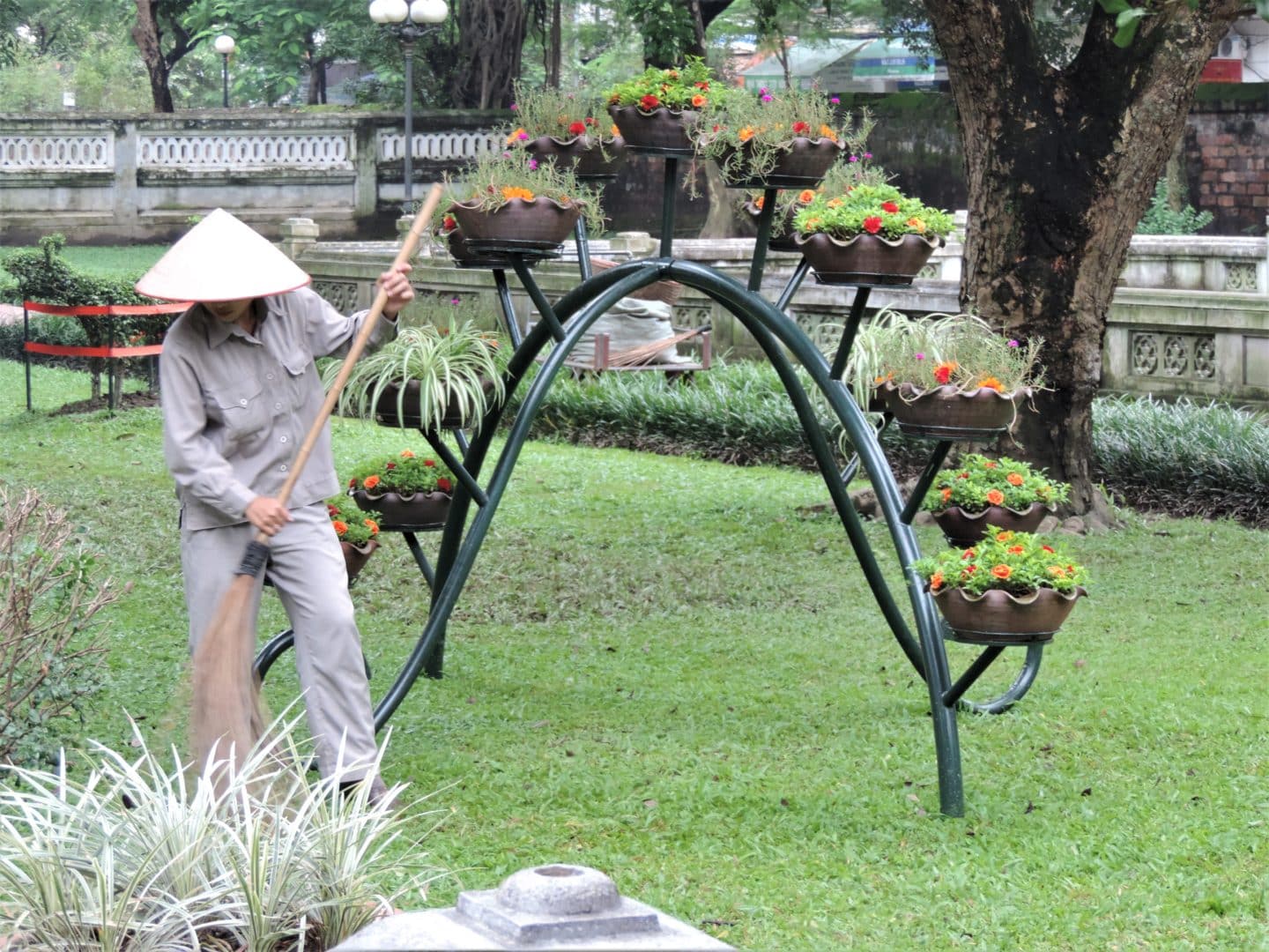 A gardener in the Literature Museum in Vietnam sweeping leaves in the courtyard area. She is dressed in a grey uniform and is holding a bamboo brush. There are floral baskets in the background in Hanoi.