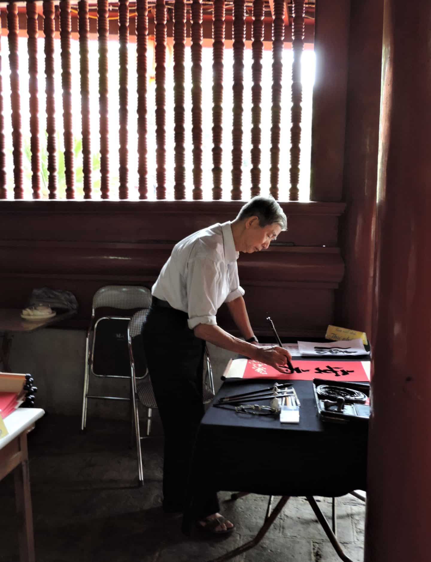 A local man writing with ink on a red piece of paper at the Literature Museum in Hanoi. He is wearing a white shirt adn dark trousers.