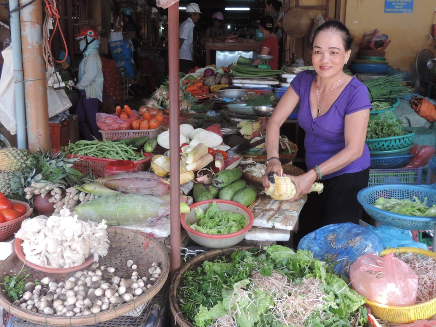 Market trader lady in Hoi An selling vegetables at the main market and cutting some root veg. She is wearing a purple t-shirt and black skirt