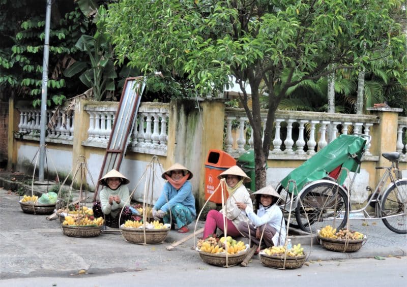 Fruit seller ladies sitting outside in Hoi An town with their baskets full of bananas and all wearing traditional hats.