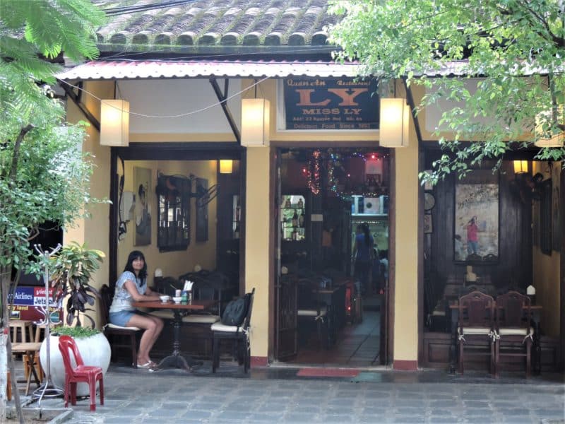 Bejal sitting outside Miss Ly in Hoi An centre waiting for dinner. Bejal is wearing denim shorts and a white and blue floaty blouse.