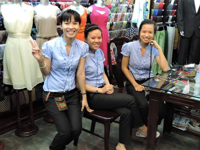 3 Tailor ladies in Hoi An wearing blue shirts and black trousers with mannequins in the background