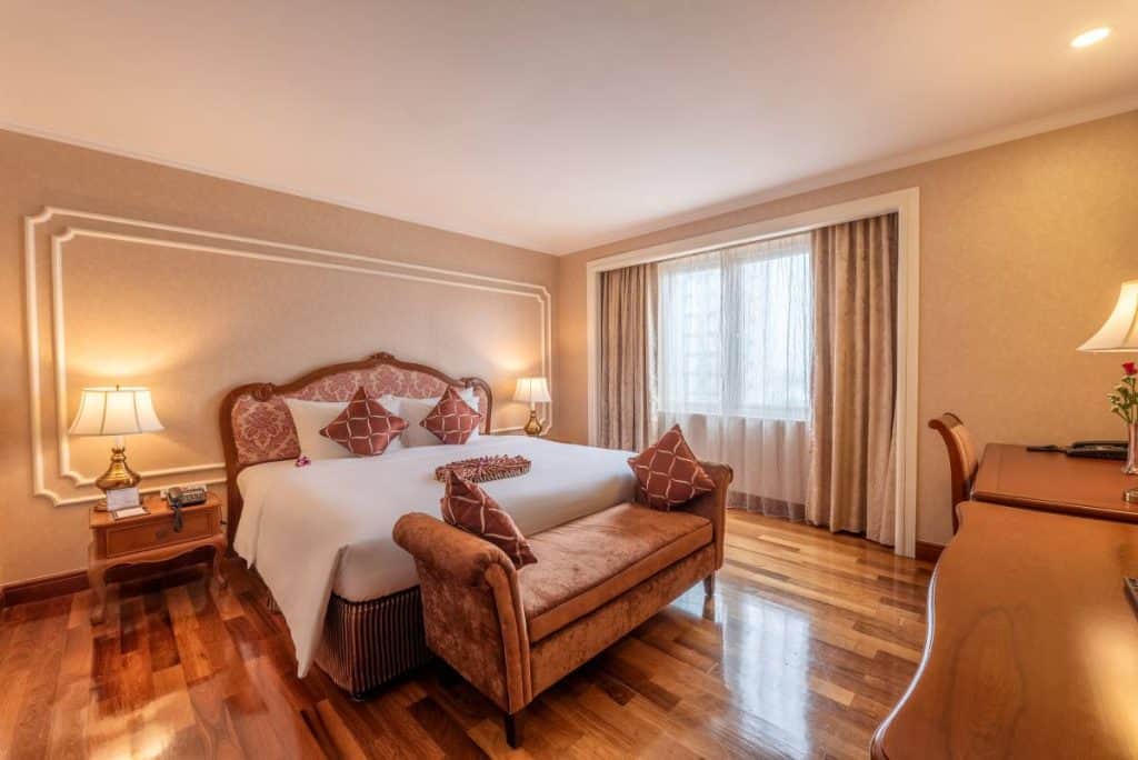 Grand Hotel Saigon Bedroom with ornate bed decorated with rust coloured cushions and a headboard with white bedding. 