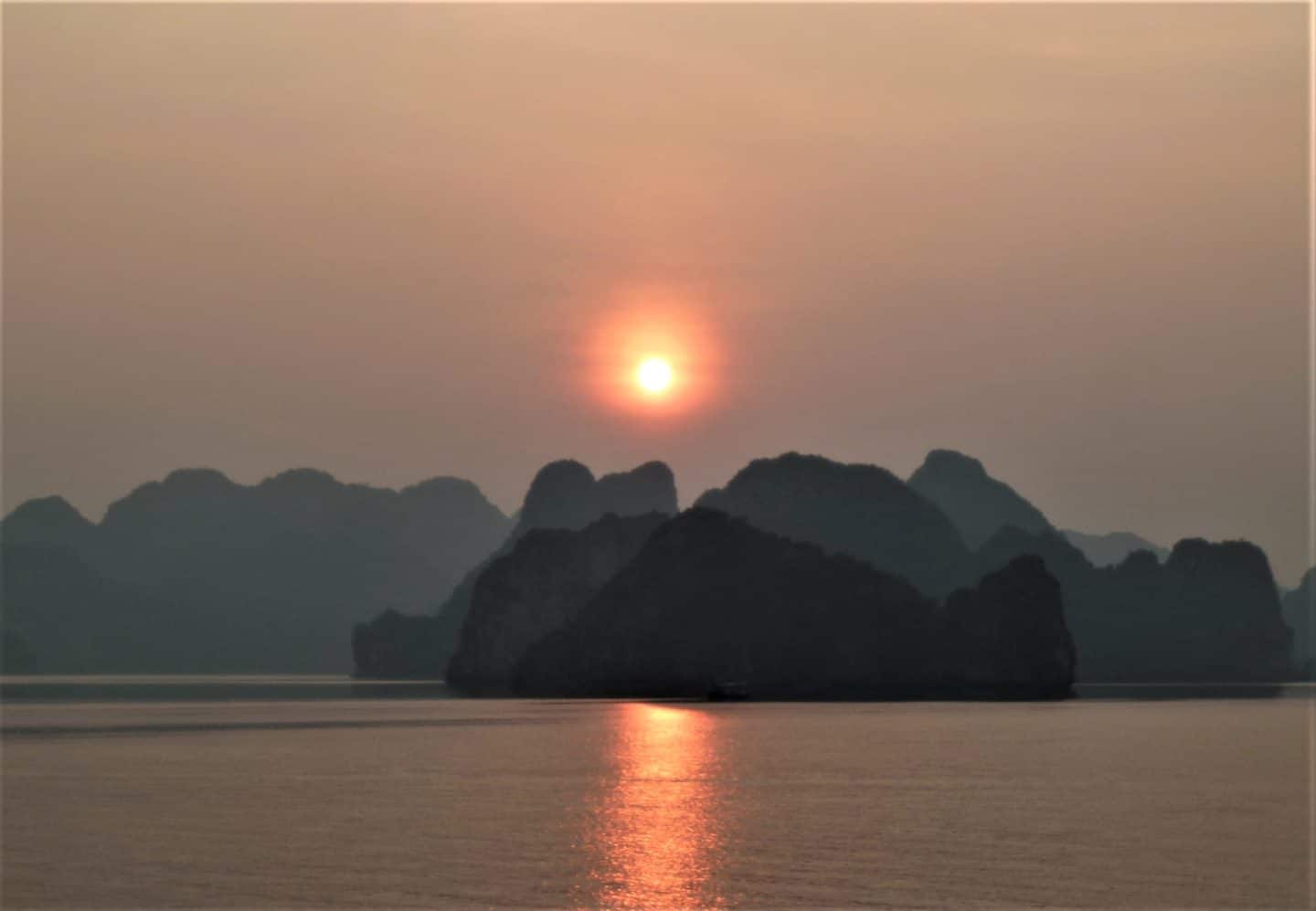 Sunrise on Halong Bay peeping through the limestone rock formations and a orange, red reflection in the sea.