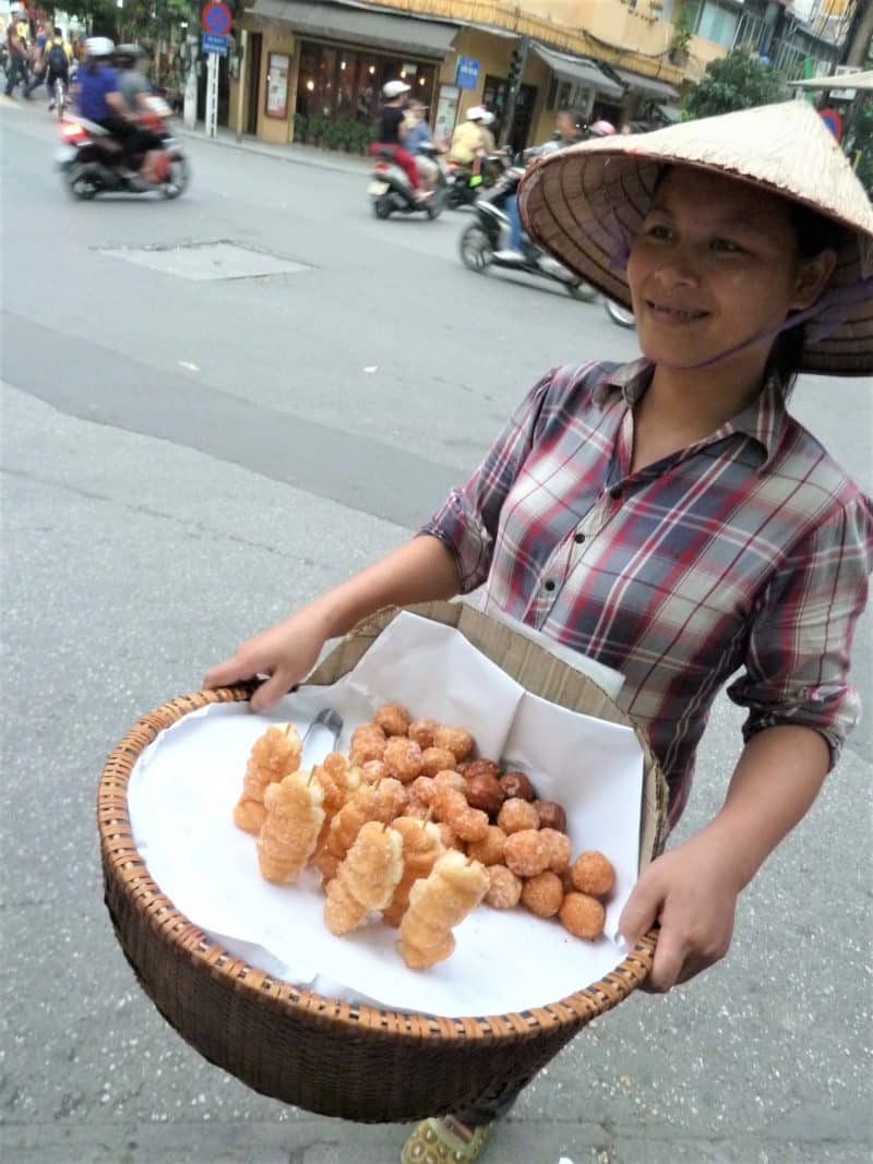 14 days in Vietnam: Lady in Hanoi selling doughnuts wearing a traditional hat, checked top and carrying doughnuts in a wicker basket