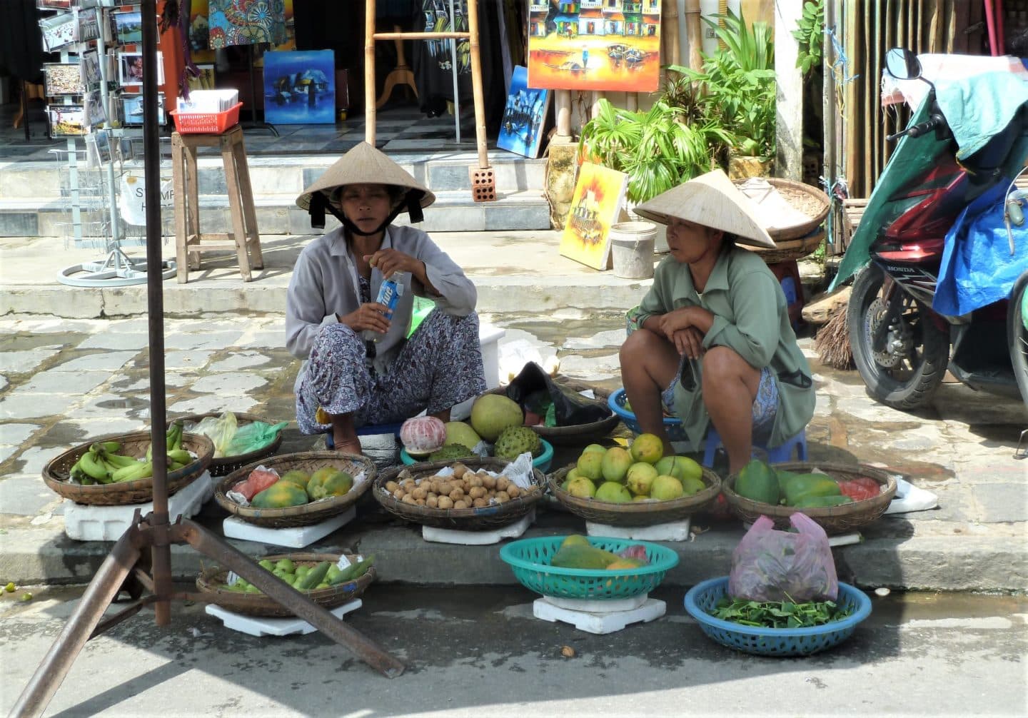 2 women in Hoi An selling fruit and vegetables. They are sitting on stools on the floor with the traditional Vietnamese hats on.