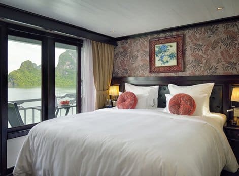 A deluxe cabin suite abourd the Paradise Luxury cruise ship. There are french doors that open up onto a balcony with a table and chair set. The bed has white linen with pink floral circular scatter cushions.