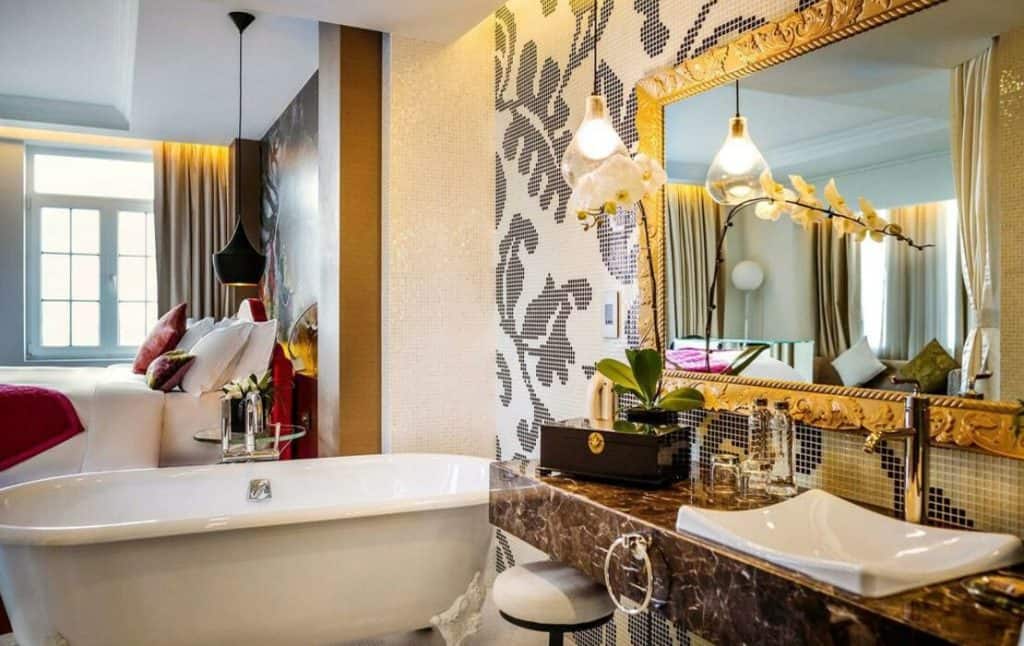 Hotel D'Lopera, Hanoi. A deluxe room with an ornate gold mirror and a vanity basin. Reflected in the mirror is a white orchid and the bedroom area with a white roll top bath.