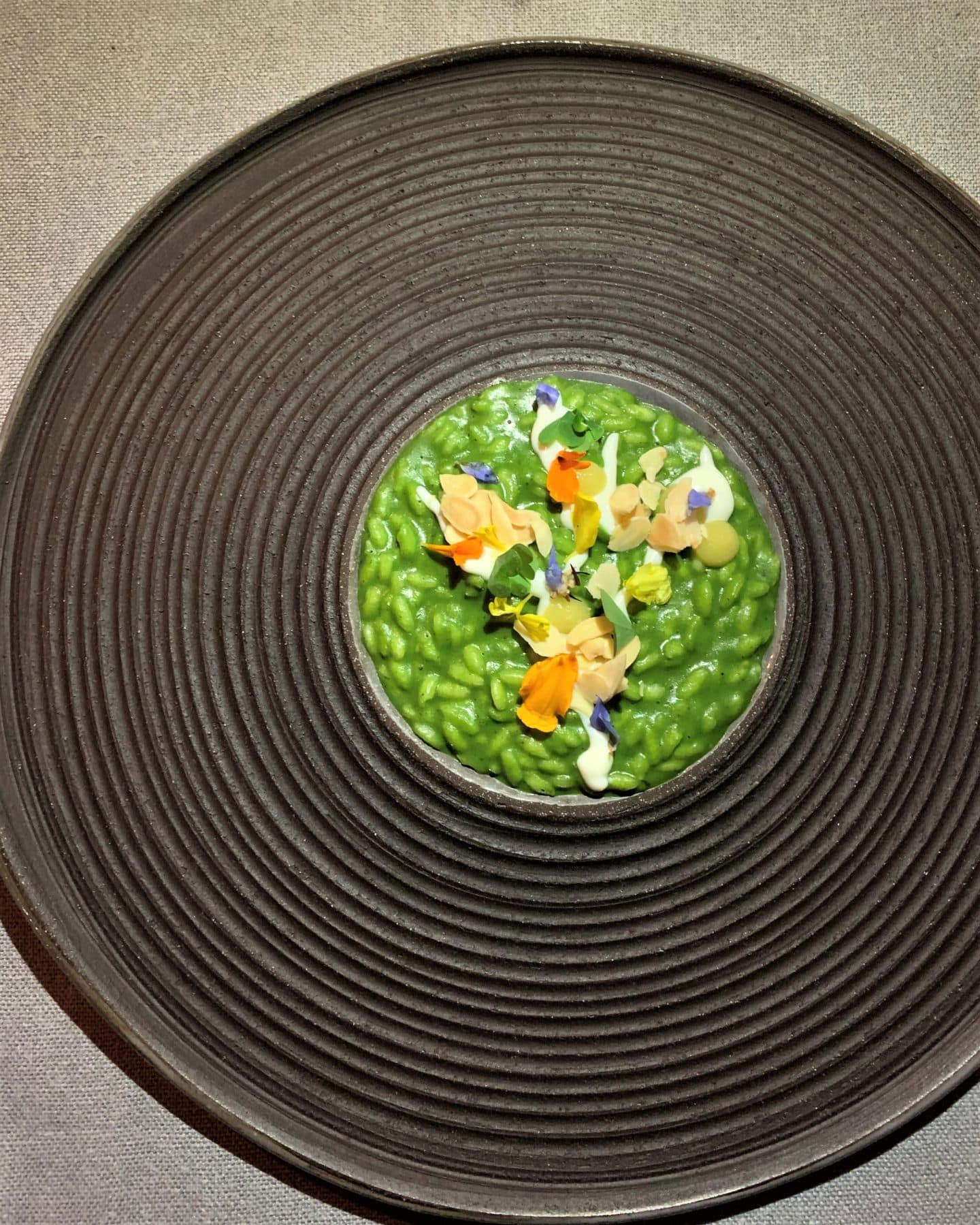 A dark brown circular plate with a green risotto in the centre wopped with edible yellow and orange flowers at la Bottego del Buon Caffe.