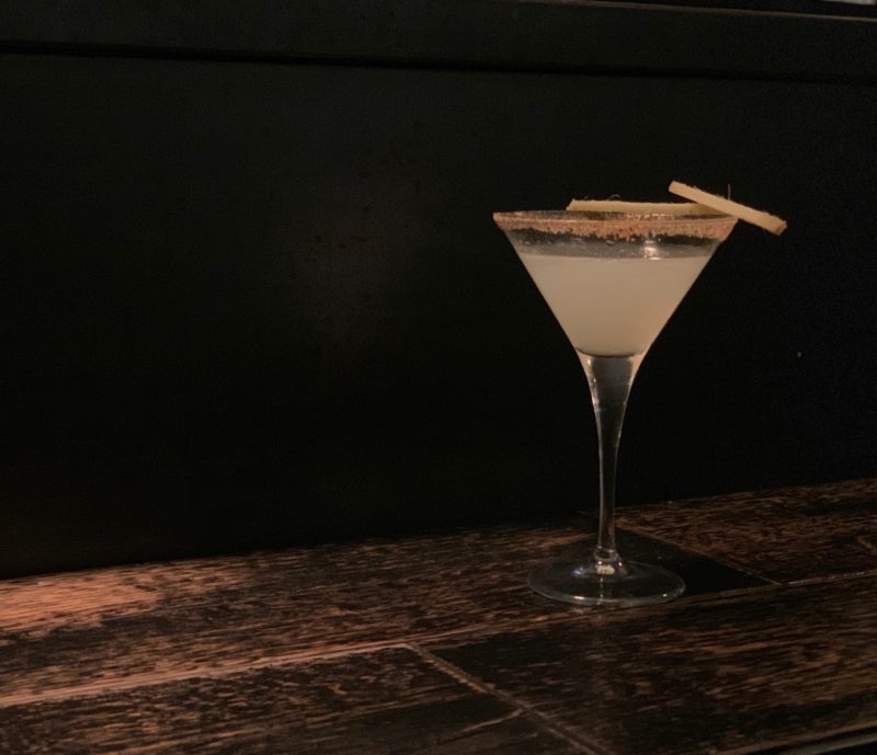 A Mezcal Gingerita cocktail at Soul Kitchen in a Martini glass with a black background adn wooden surface.