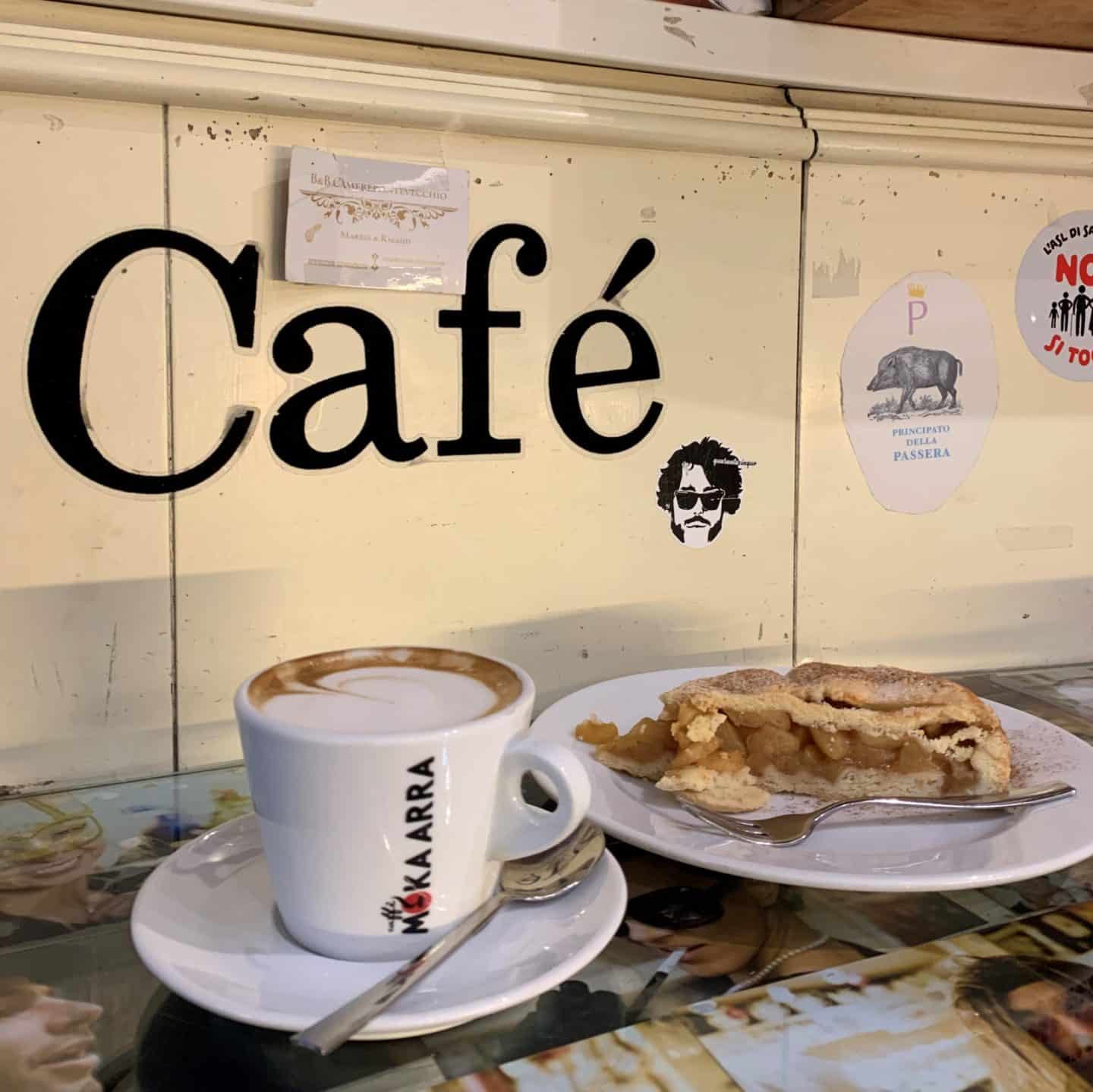 Vegetarian food in Florence. The interior of Caffè degli Artigiani cream walls adn cafe written on it. On a ledge there's a cappuccino and an apple tart cake.