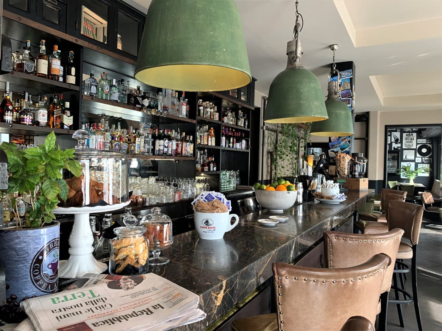 The interiors of La Drogheria in Florence with green lampshade bar lights and a black marble bar with cream studded seats