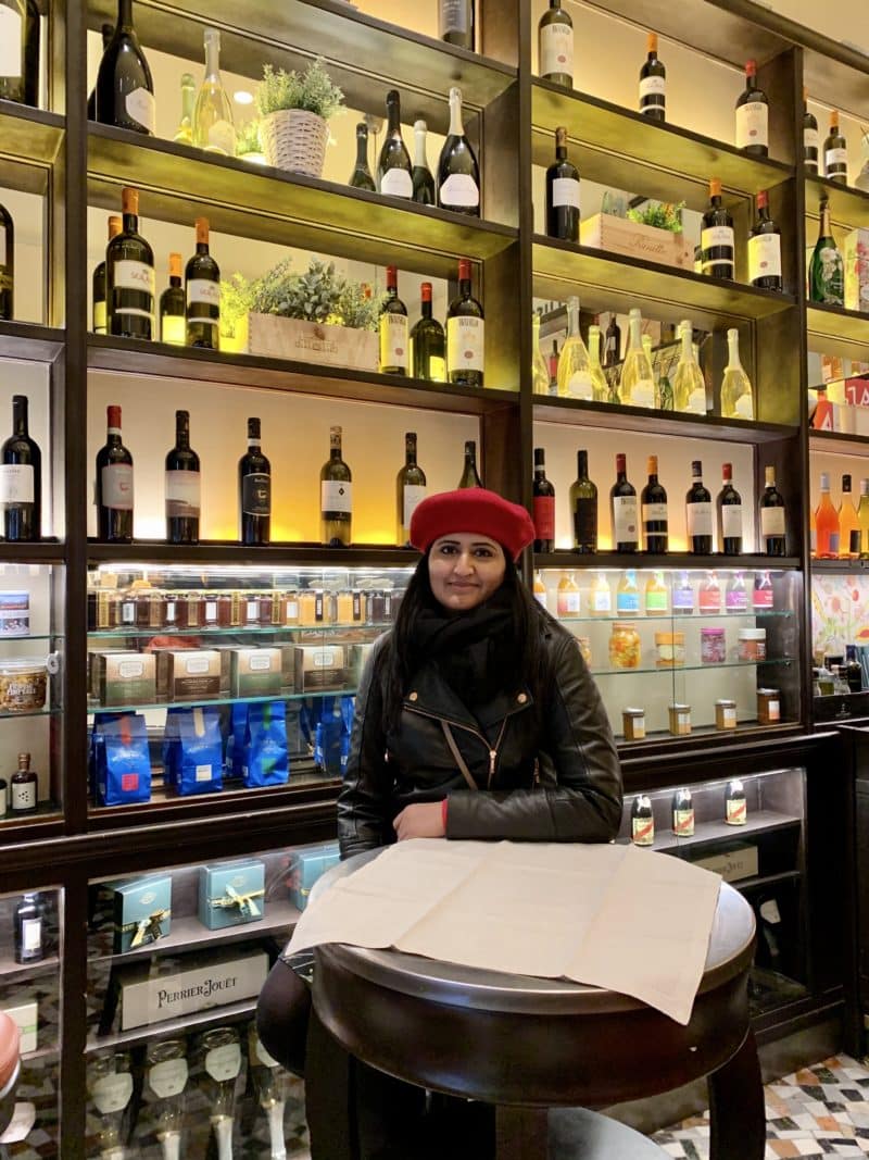 Bejal sat at bistro table The background has jars of truffles and conserves. Bejal is wearing a red berey and black leather jacket.