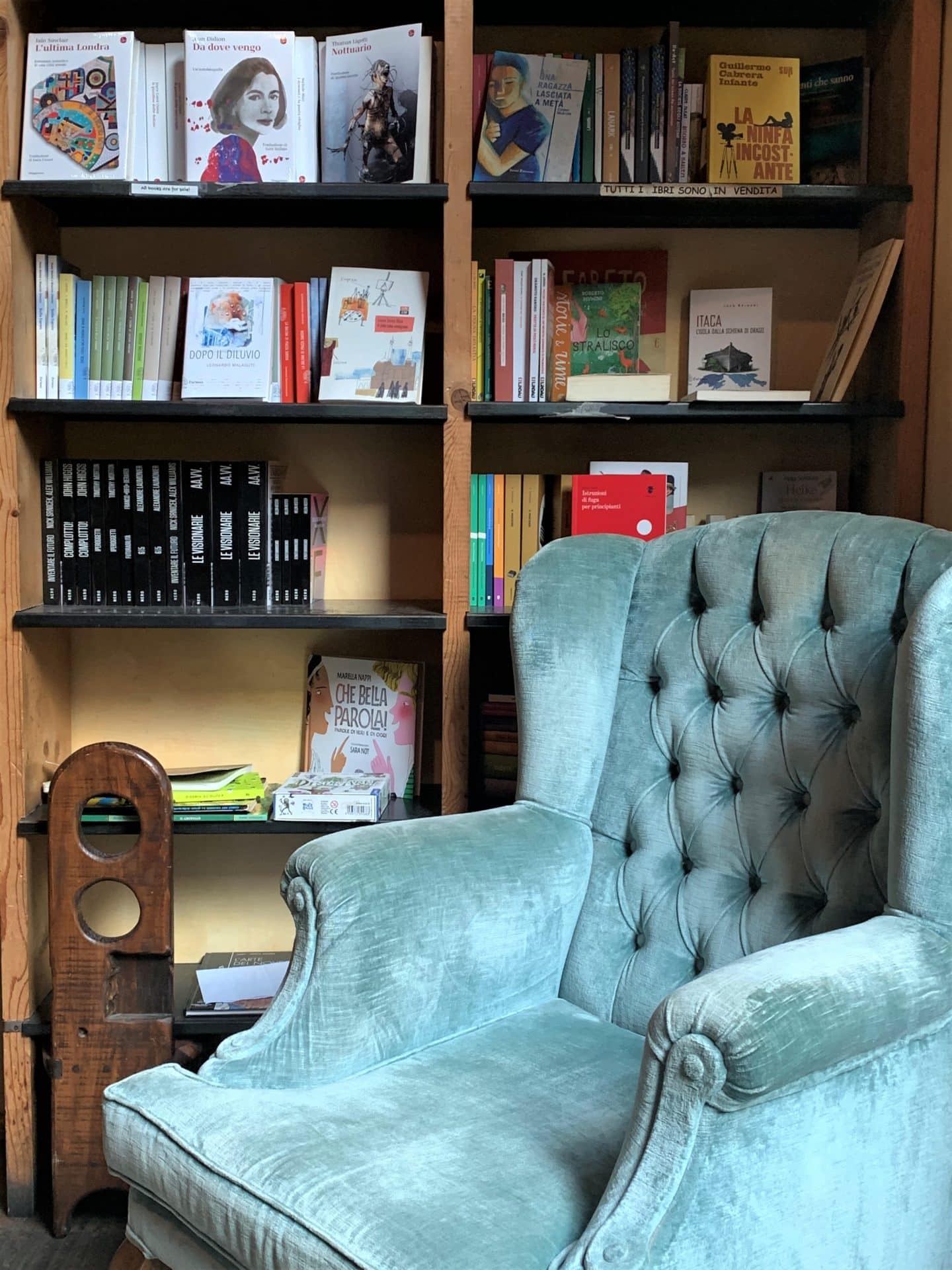 Libreria Cafè La Cité with a pale blue armchair with a buttoned back and a bookcase containing books scattered throughout.