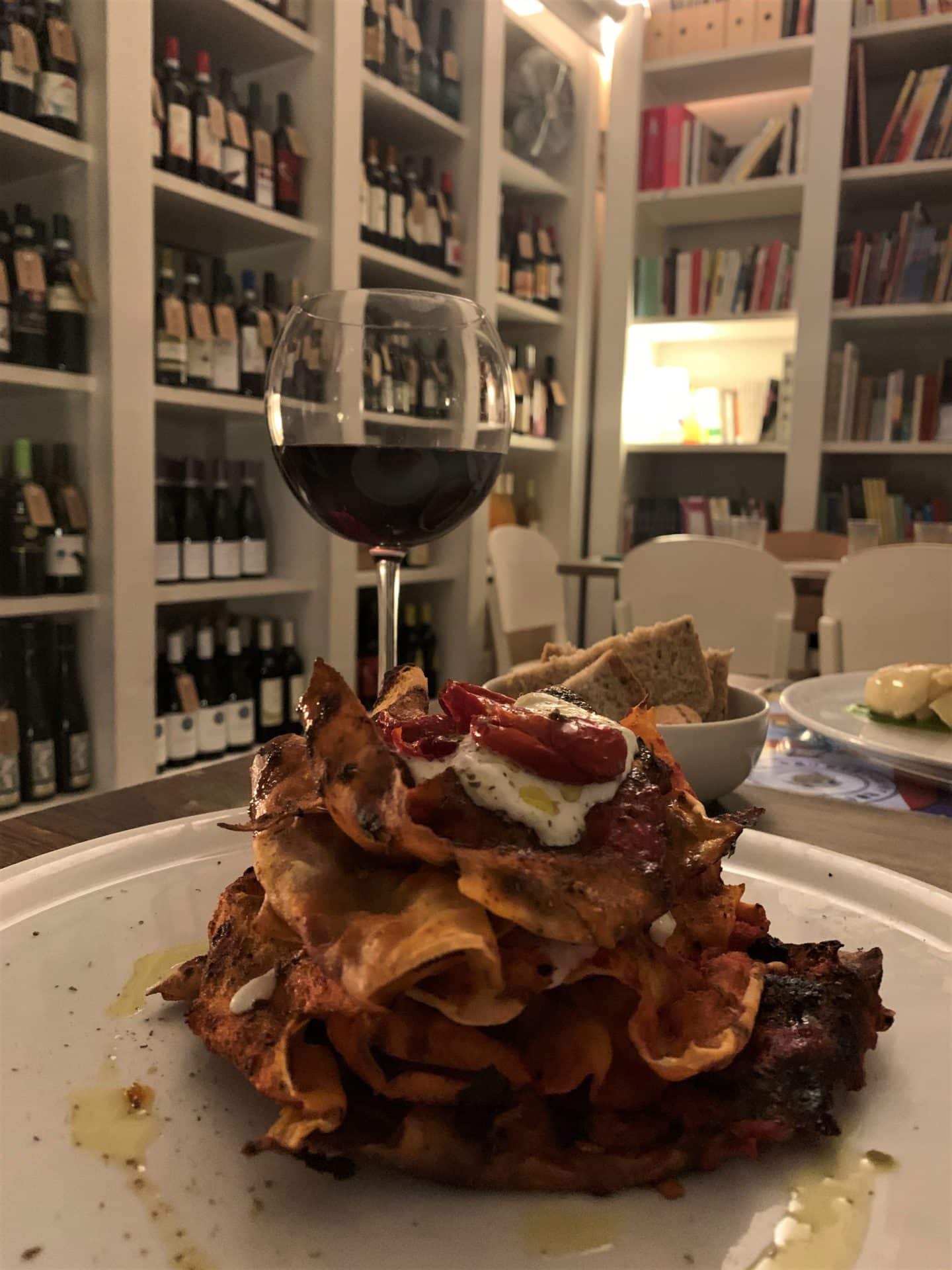 The interior book cases in BRAC with wine bottles and books and dining tables. A glass of red wine and pasta covered in tomato sauce.