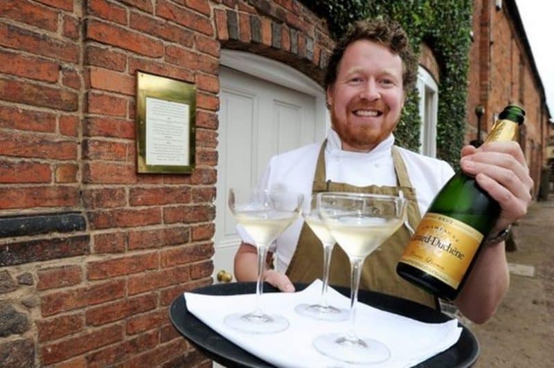 Michelin Star dining at John's House in Leicestershire: John holding a bottle of champagne and 3 glasses full of champagne, outside his restaurant