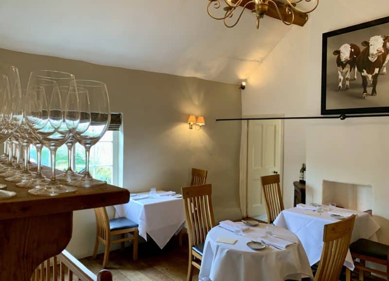 estaurant area upstairs at John's House with tables set up and a shelf of glasses. Photos of cows on the walls with a gold chandelier in the middle.