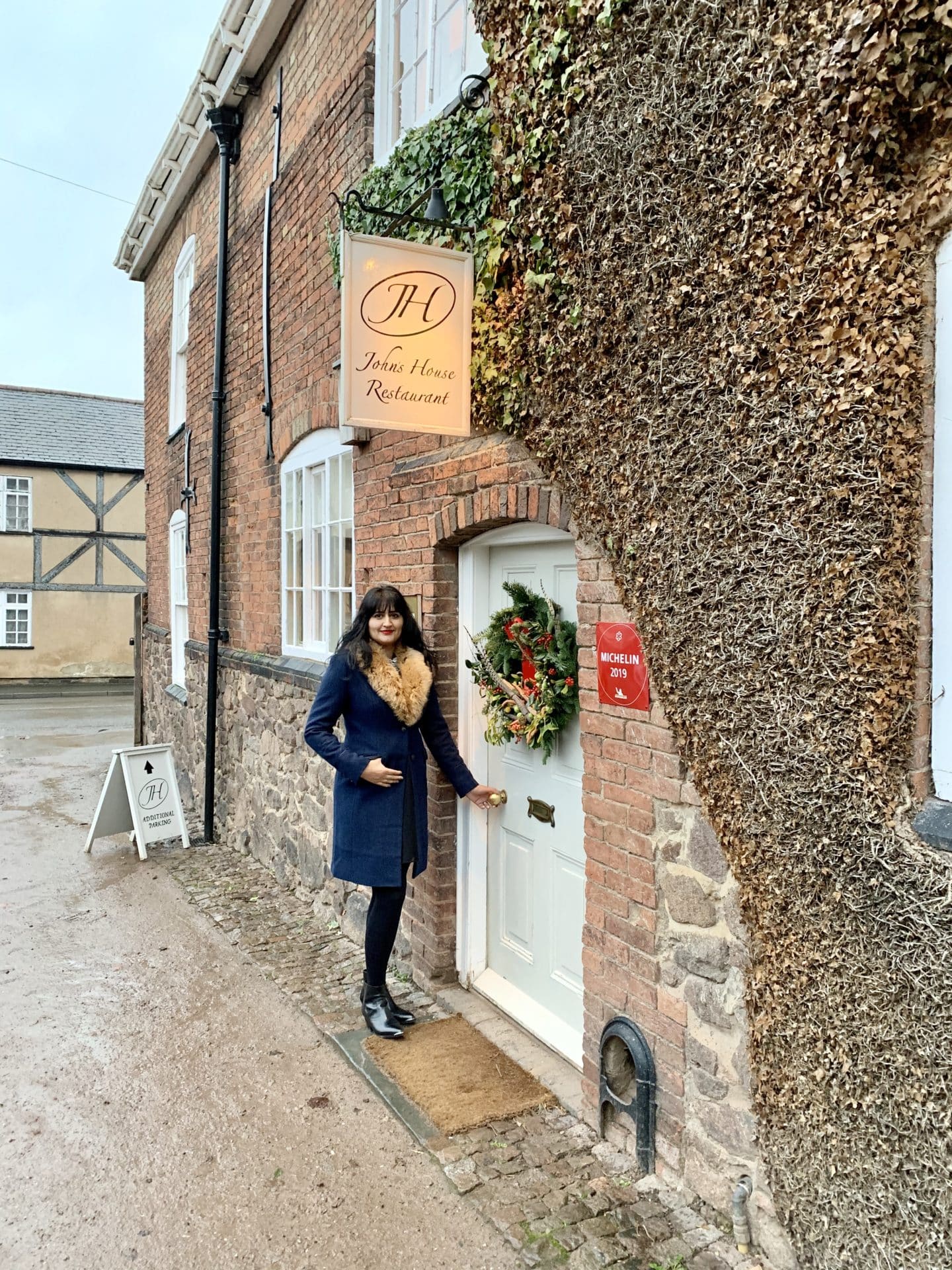 The exterior of John's House restaurant with white window frames and a white door which has a Christmas wreath. The building is a mixture of brick and stone. Bejal standing outside wearing a navy wool coat with faux fur collar.