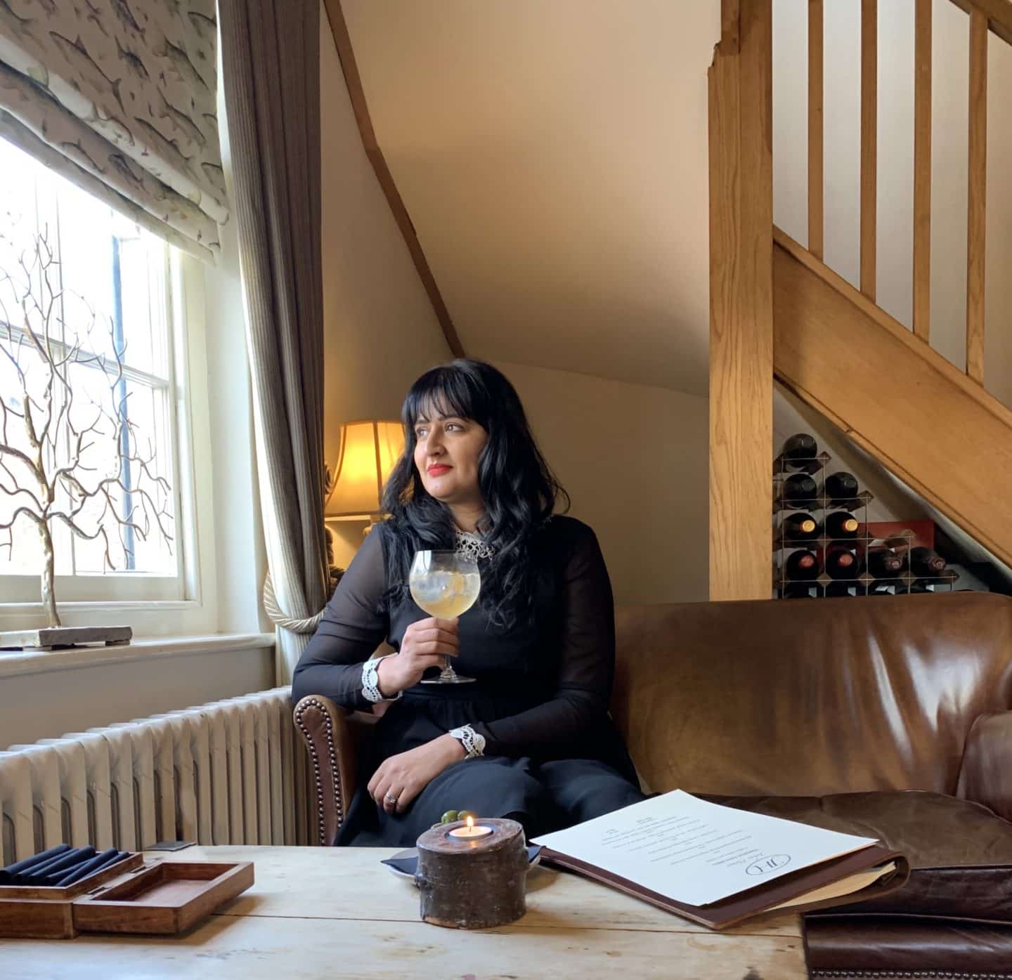 Michelin Star dining at John's House in Leicestershire: Bejal holding a gin and tonic whilst looking out of the window, wearing a black dress in the lounge