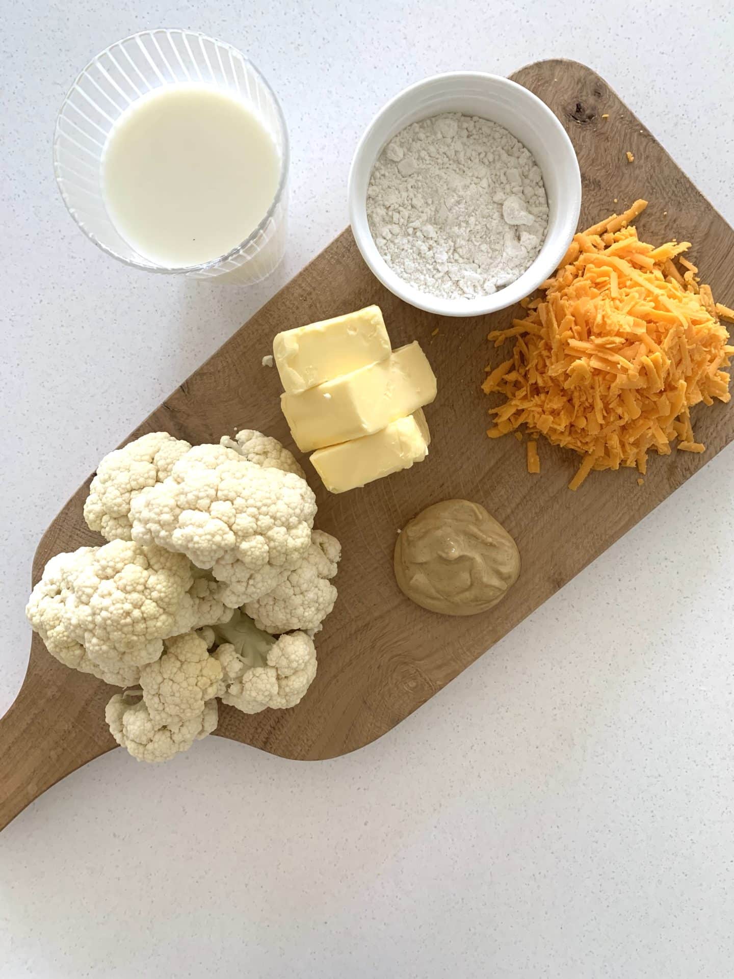 ingredients to make cauliflower cheese on a chopping board, milk, cauliflower, mustard, grated Red Leicester, plain flour and butter