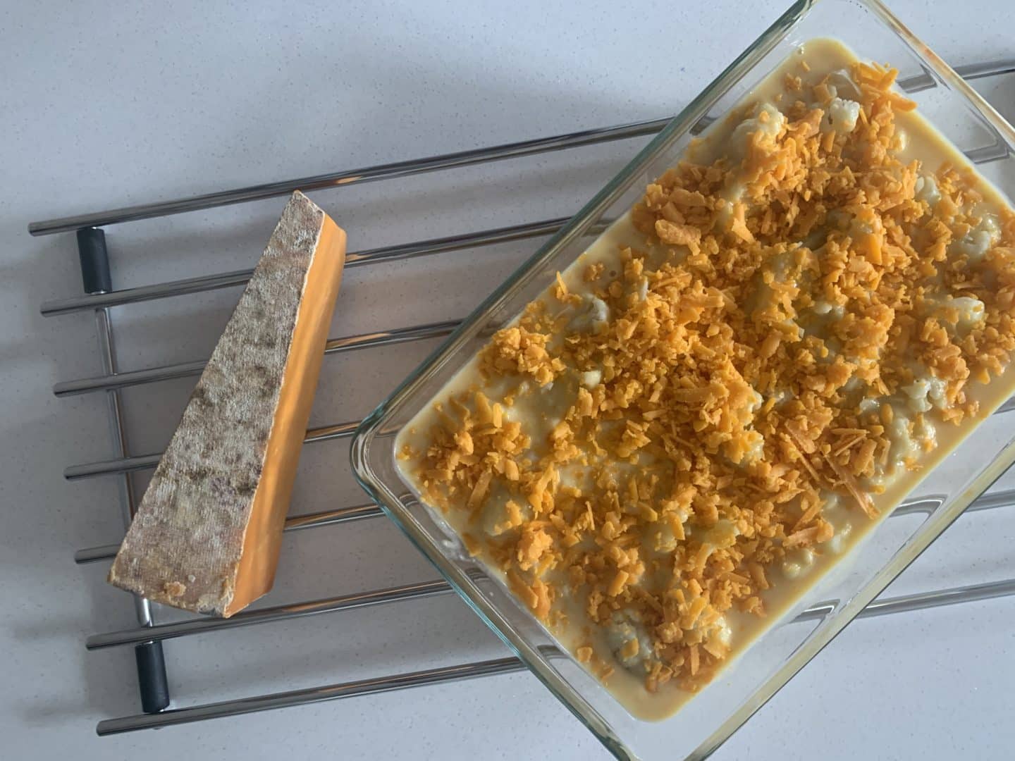 Large dish with cauliflower cheese with a slice of Red Leicester cheese on teh side on a wire rack