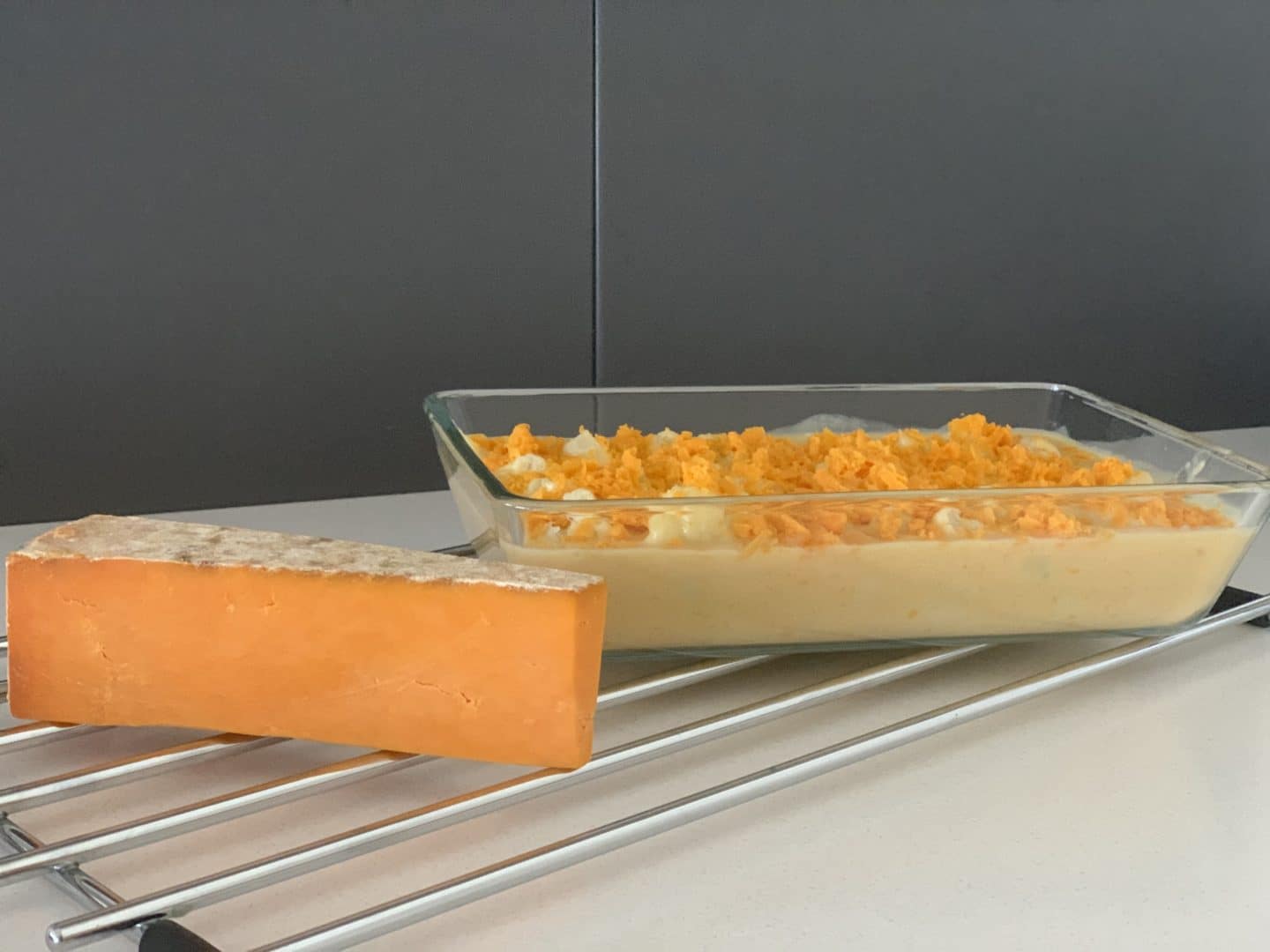 Large dish with cauliflower cheese with a slice of Red Leicester cheese on teh side on a wire rack (side view)