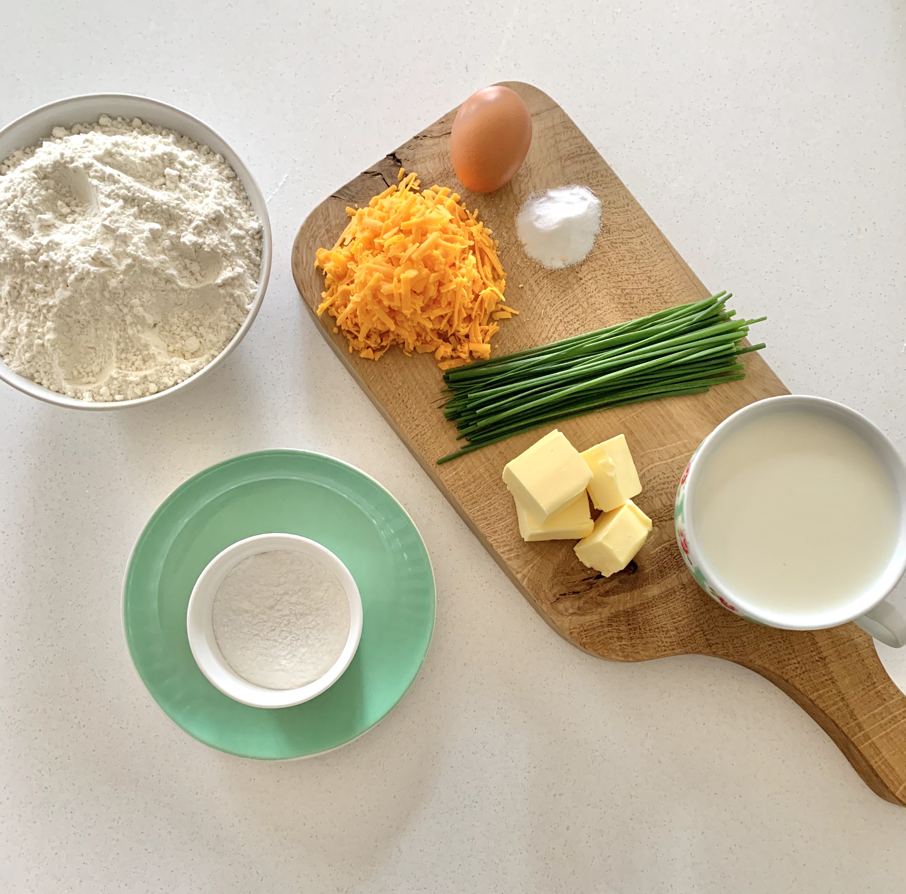 A chopping board with chives, Red Leicester grated cheese, an egg, butter pieces, milk and plain flour and baking powder