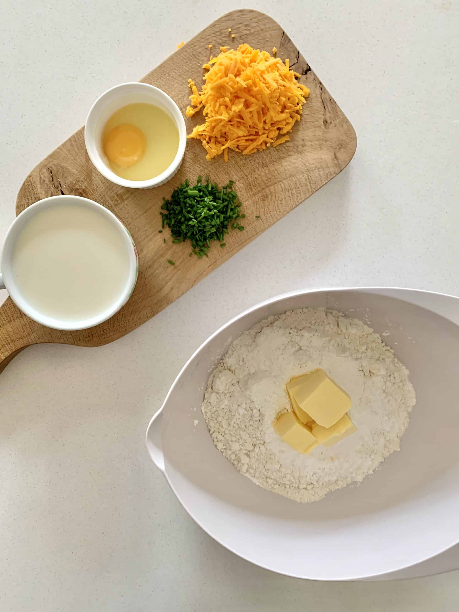 A chopping board with chives, Red Leicester grated cheese, an egg, butter pieces, milk and plain flour and baking powder. Butter pieces and flour are in a white bowl.