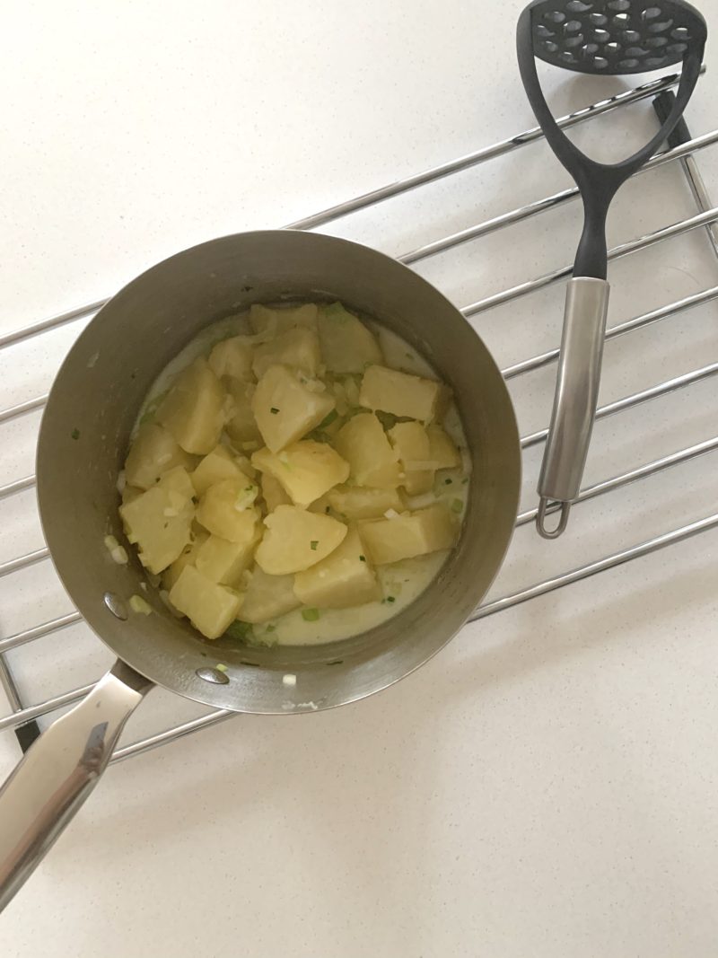 Sauce pan with chopped boiled potatoes, butter and milk, ready for mashing. A silver masher is placed on the side
