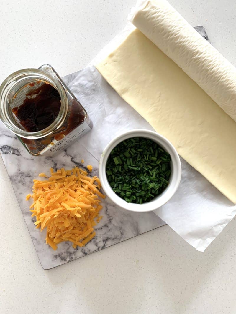 Roll of pastry, cut chives, grated Red Leicester cheese and red onion chutney on a marble mat