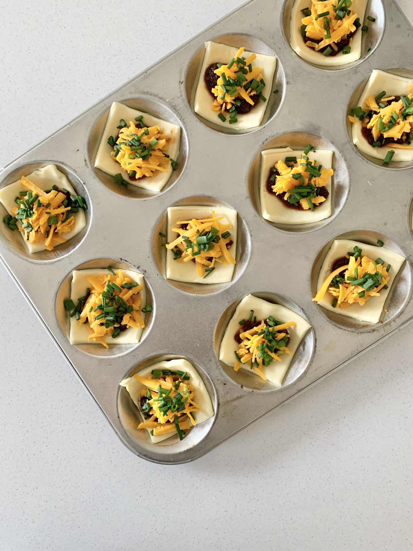 Tray with pastry squares, topped with red onion chutney, grated Red Leicester cheese and chives ready to be baked.