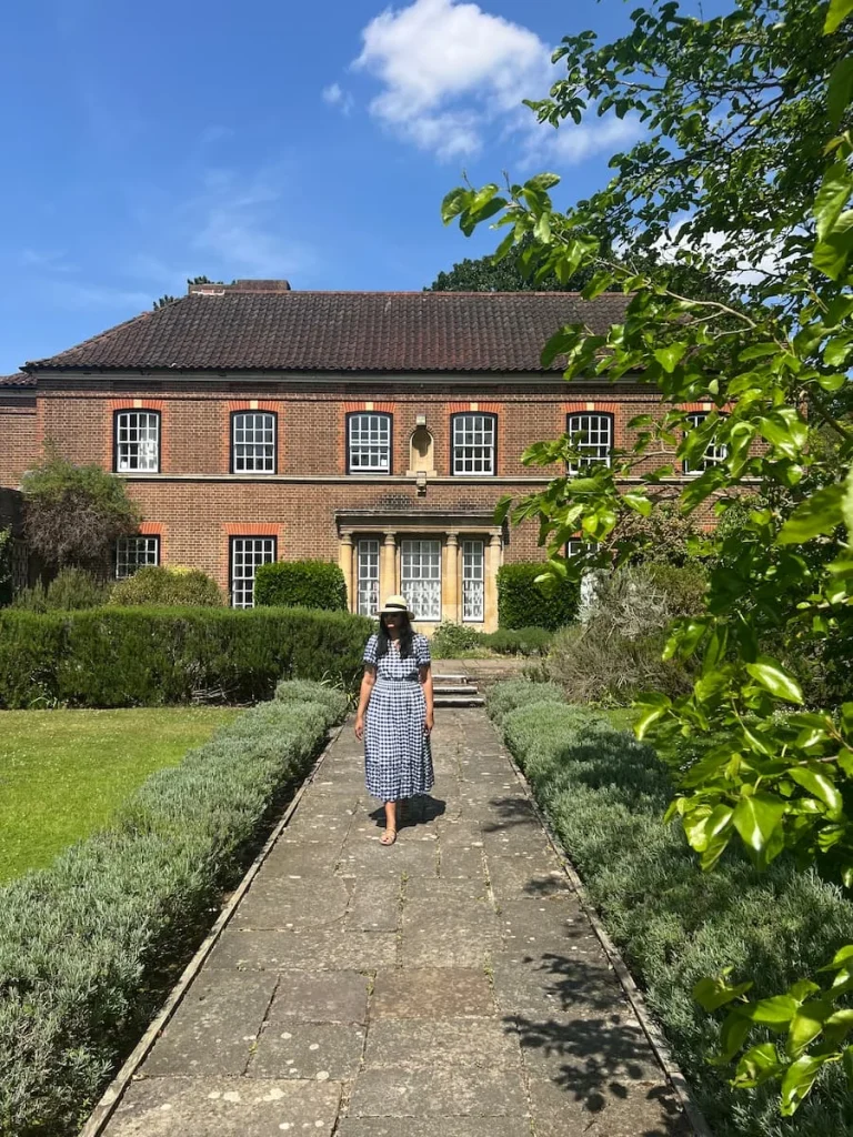 Bejal walking down a tree lined path wearing a blue and white checked dress and a hat with the manor house in the background