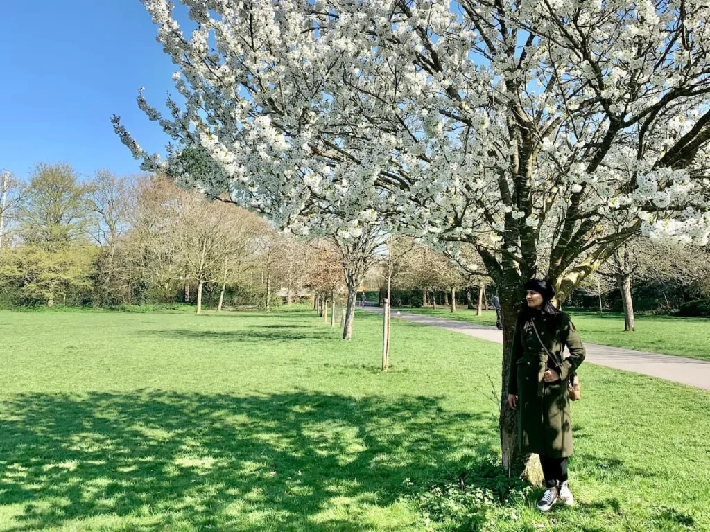 Bejal on a cool spring day wearing a coat and trainers standing under a white blossom tree, Leicester