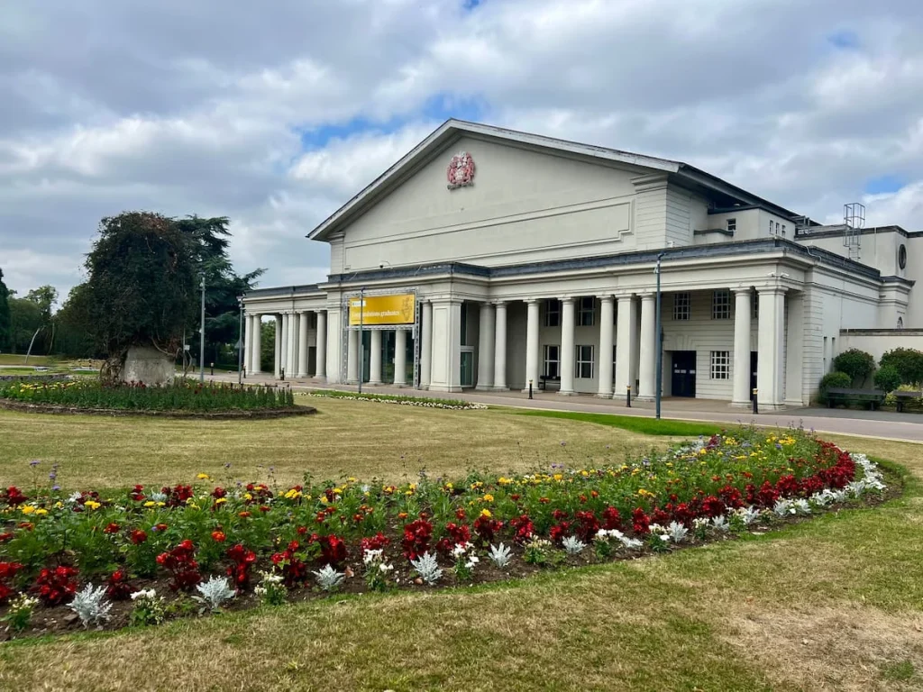 Exterior of De Montfort Hall in Leicester. Multicoloured flower beds at the front which contrast the white and cream exterior of the building work with big white pillars at the entrance.