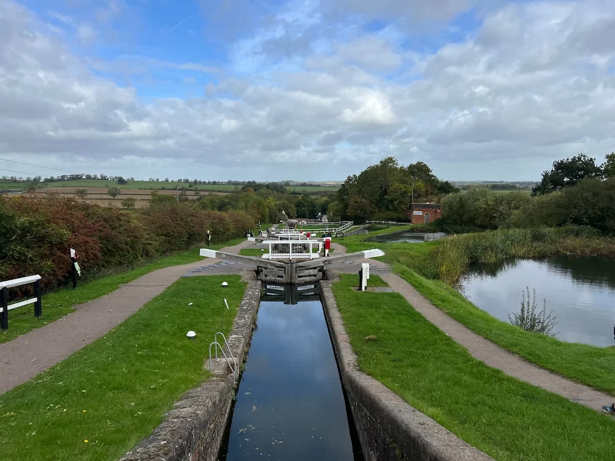 Foxton Locks from Top Lock, Leicestershire. The locks are in the centre with the green grass adn foliage surrounding them with a blue sky with white fluffy clouds. 
